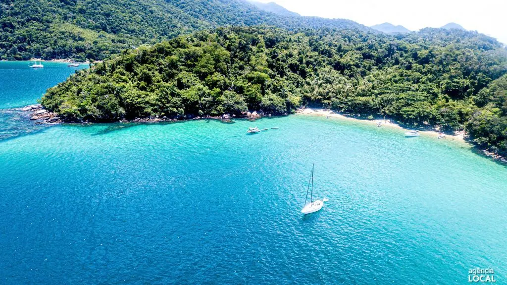 Enseada de água azul-turquesa com veleiro, pequena praia e encosta coberta de mata na Ilha Grande