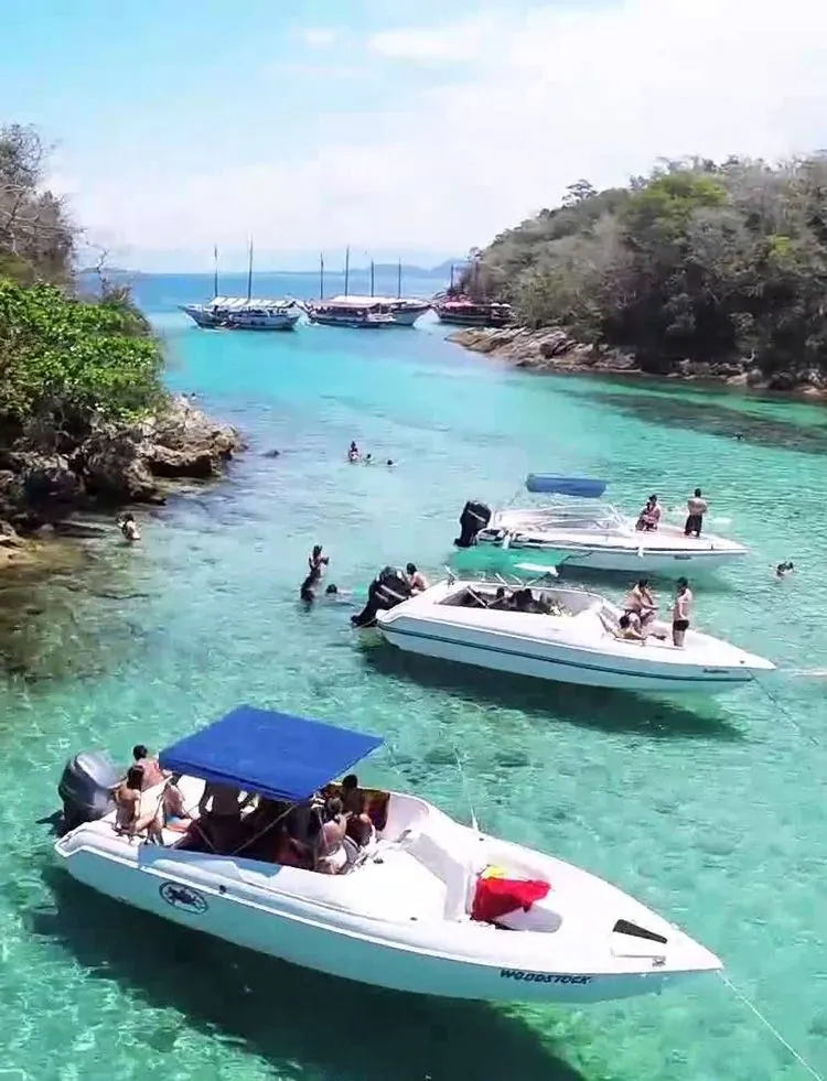 Lanchas e escunas ancoradas em canal de água cristalina entre costões, com pessoas nadando - passeios de lancha na Ilha Grande