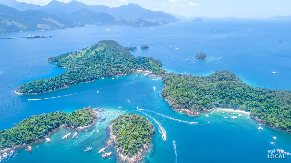 Ilhas cobertas de Mata Atlântica e águas azul-turquesa com escunas e lanchas, com as serras do continente ao fundo - Ilha Grande, RJ